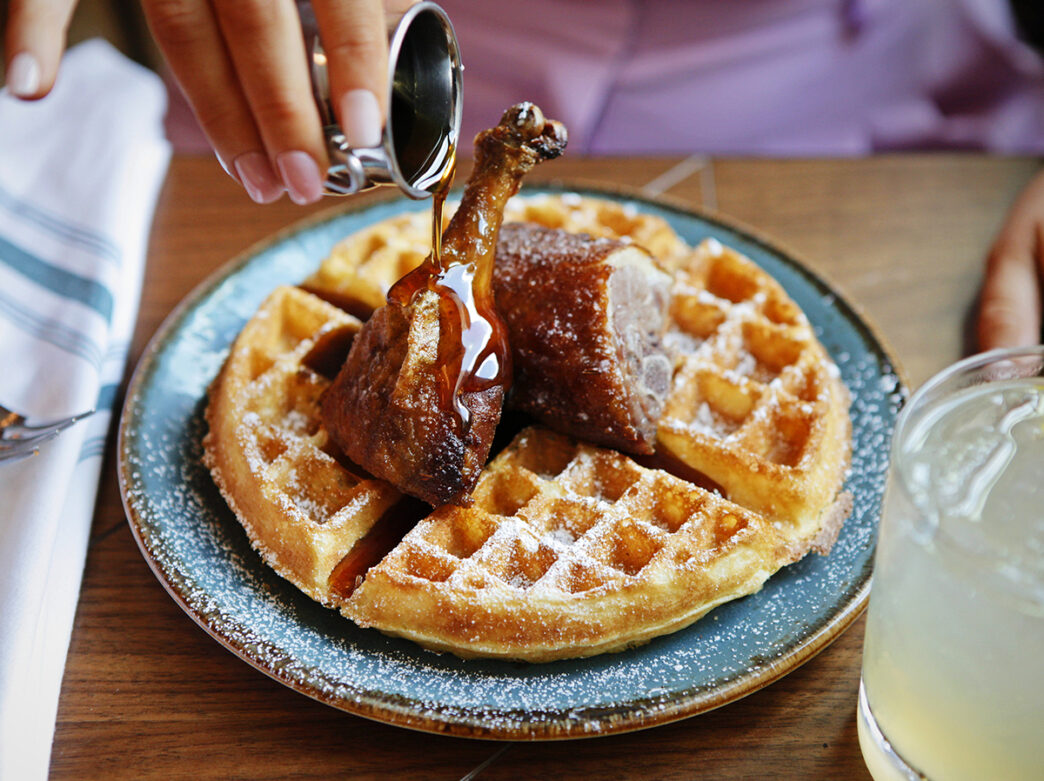 A close up image of a Belgium waffle topped with crispy fried chicken as syrup is poured over the dish.