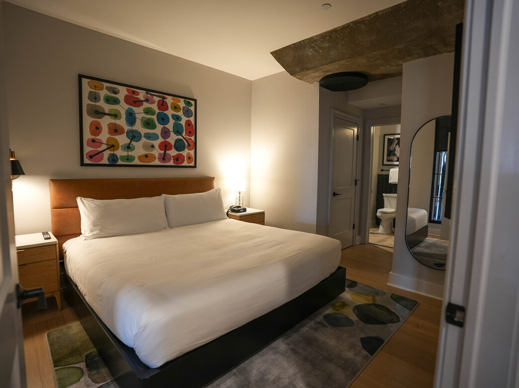 A modern guest room featuring a king-sized been with white linens, a tan leather headboard, colorful abstract artwork and contemporary furnishings.