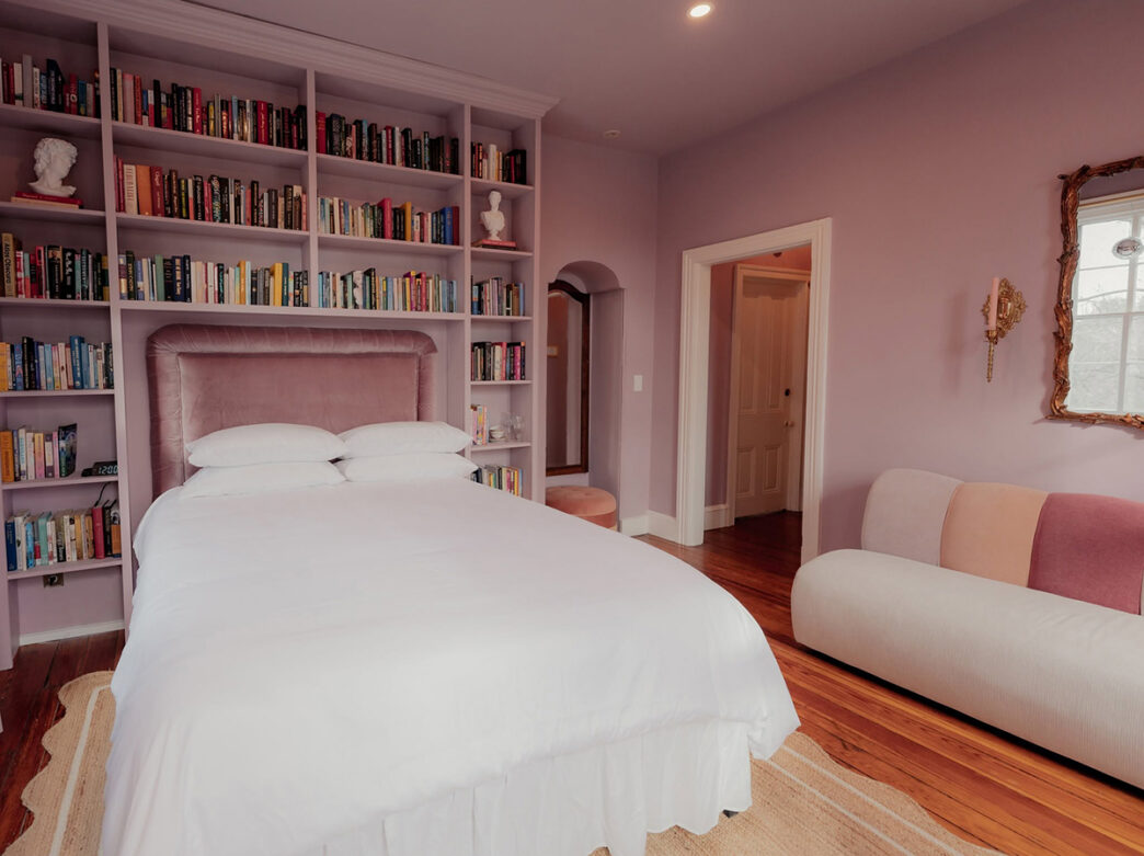 A hotel room featuring soft lavender walls, floor-to-ceiling bookshelves surrounded a plush bed with white linens, and a cozy pastel-colored sofa.