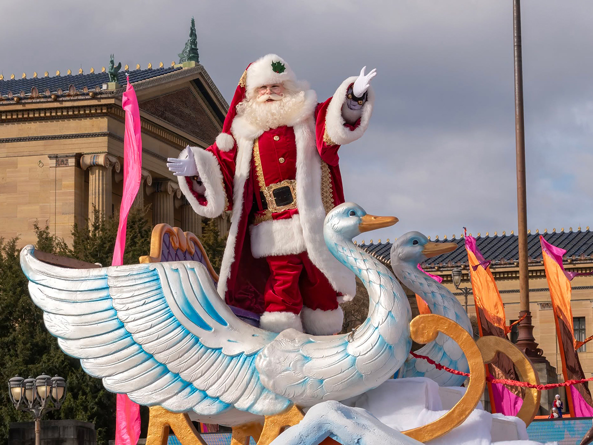Claus waves to spectators from a festive float featuring oversized white swans during Philadelphia's 6abc Dunkin' Thanksgiving Day Parade, with the Philadelphia Museum of Art in the background.