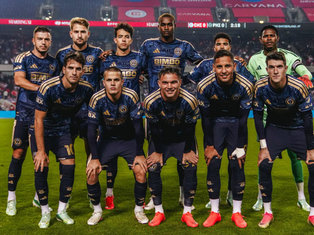 Un grupo de once futbolistas profesionales posa para una foto de equipo en el campo de fútbol antes de un partido; todos llevan uniformes azul marino y dorados, y el portero lleva una camiseta verde.