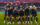 A group of eleven professional soccer players pose for a team photo on the soccer field before a match, wearing navy blue and golden uniforms with the goalkeeper in a green jersey.