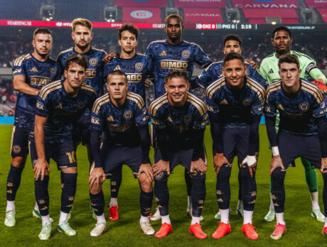A group of eleven professional soccer players pose for a team photo on the soccer field before a match, wearing navy blue and golden uniforms with the goalkeeper in a green jersey.