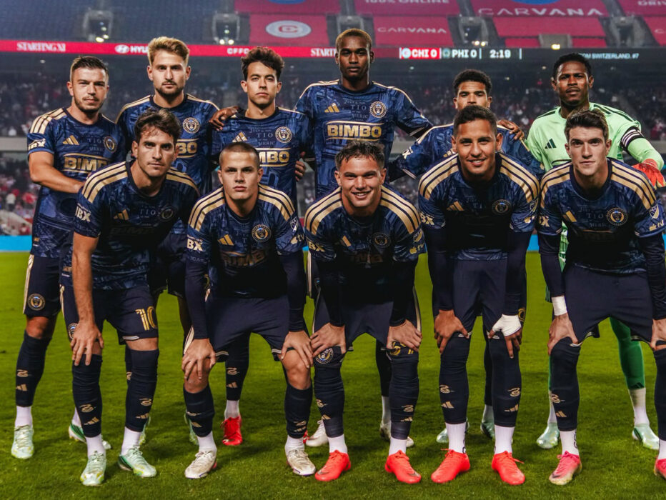 A group of eleven professional soccer players pose for a team photo on the soccer field before a match, wearing navy blue and golden uniforms with the goalkeeper in a green jersey.