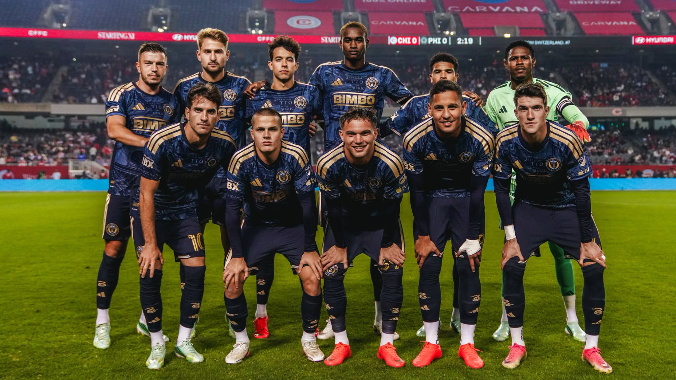 A group of eleven professional soccer players pose for a team photo on the soccer field before a match, wearing navy blue and golden uniforms with the goalkeeper in a green jersey.