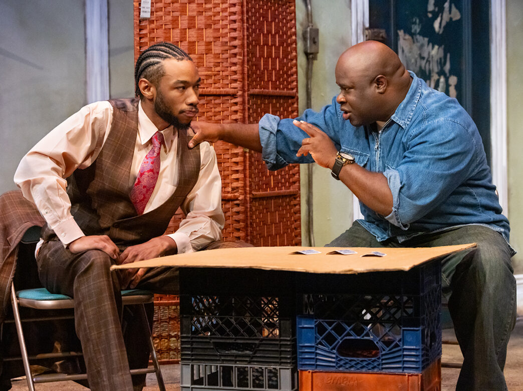 Two actors perform a scene on stage during a production of Topdog/Underdog, with one wearing a brown suit and the other a denim shirt, engaged in an intense conversation across a makeshift table.
