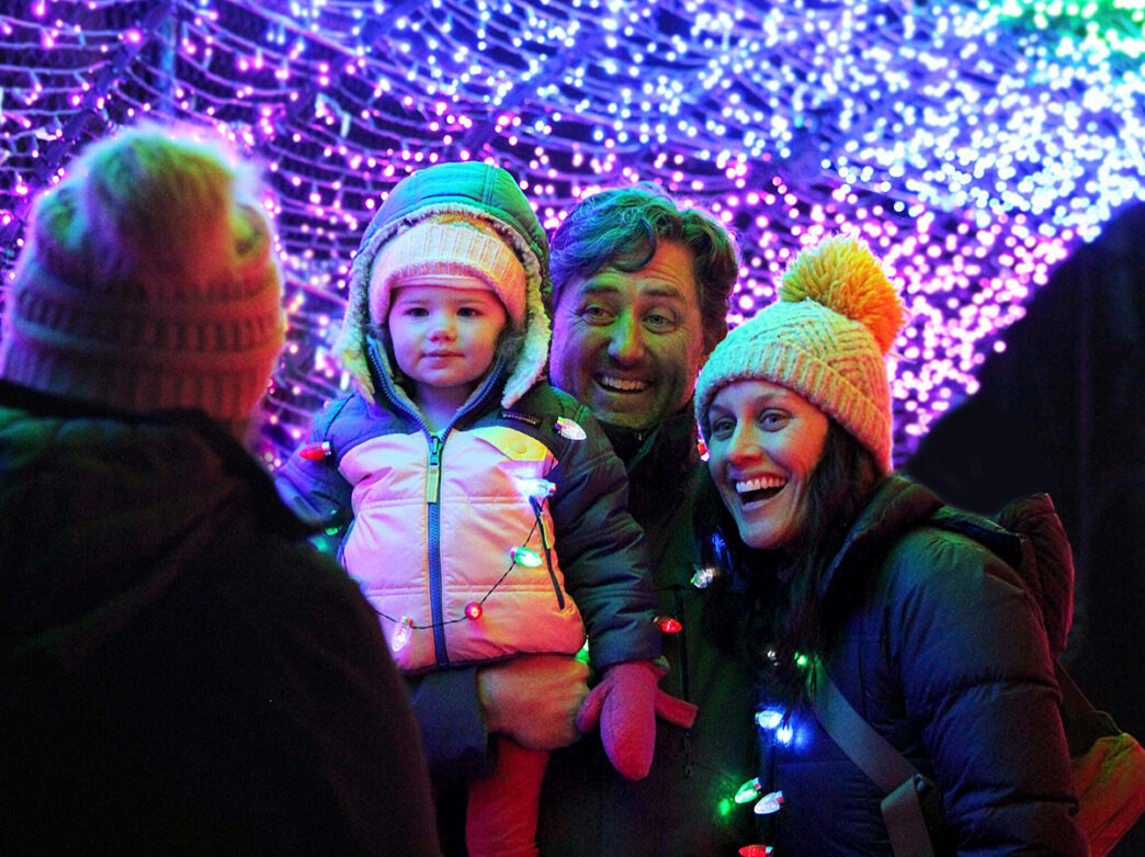 A smiling family dressed in winter coats and knit hats poses beneath a glowing canopy of purple, pink, green and blue holiday lights.