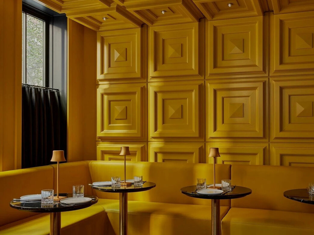 A stylish, monochromatic yellow lounge at Andra Hem featuring plush banquette seating, geometric wall paneling and black marble tables.